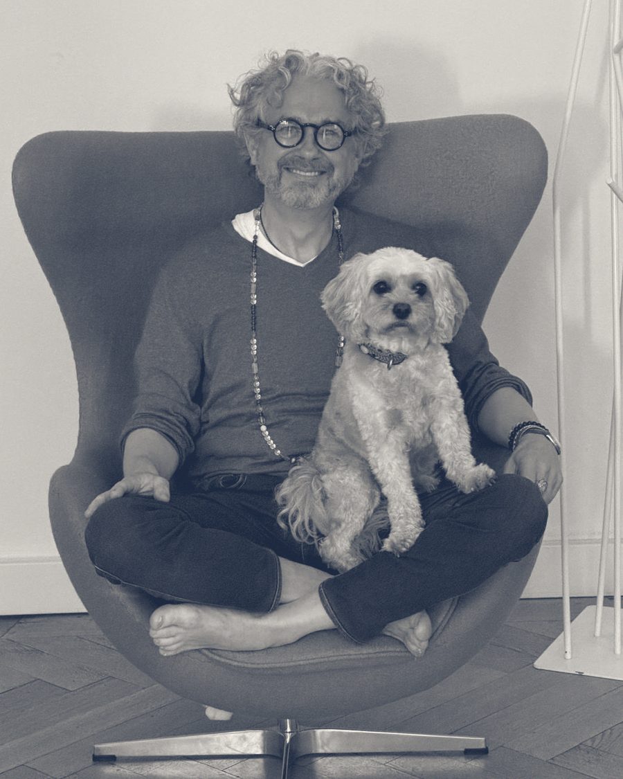 Christopher Maria Ofenstein sitzt barfuß im Schneidersitz in einem Sessel, einen kleinen hellen Hund auf dem Schoß.